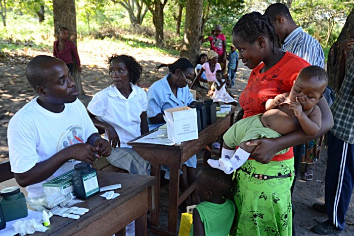 Manhica's local health workers register and track each child vaccinated at the mobile session. A mother and her child talk with a male health worker in the cool of the shade in their makeshift clinic.