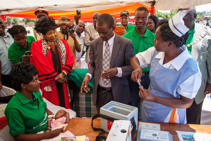 Honourable Dr Joseph Katema, Minister of Community Development, Mother and Child Health, speaks with Lisa Chinyama, the mother of the first child to be vaccinated during the ceremony, as a community health worker prepares the vaccines.