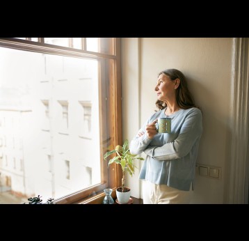 Portrait of thoughtful retired mature woman in casual clothes standing by a window drinking tea.