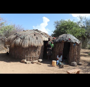 Angelina Muthoni stands outside their hut in Aremet with her mother sitting enjoyng the house's shade with her other child. Credit: Dominic Kirui
