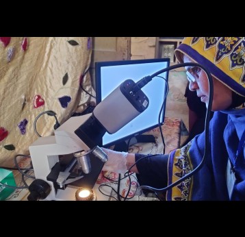 A female health worker looks through a microscope connected to a computer screen, examining a sample during a community health session