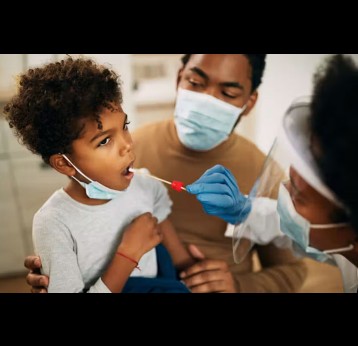 A child receives a swab to test for covid. He is being held by his father, who is wearing a face mask. A female health worker is performing the swab. She is also wearing a face mask. Credit: Drazen Zigic/ Shutterstock