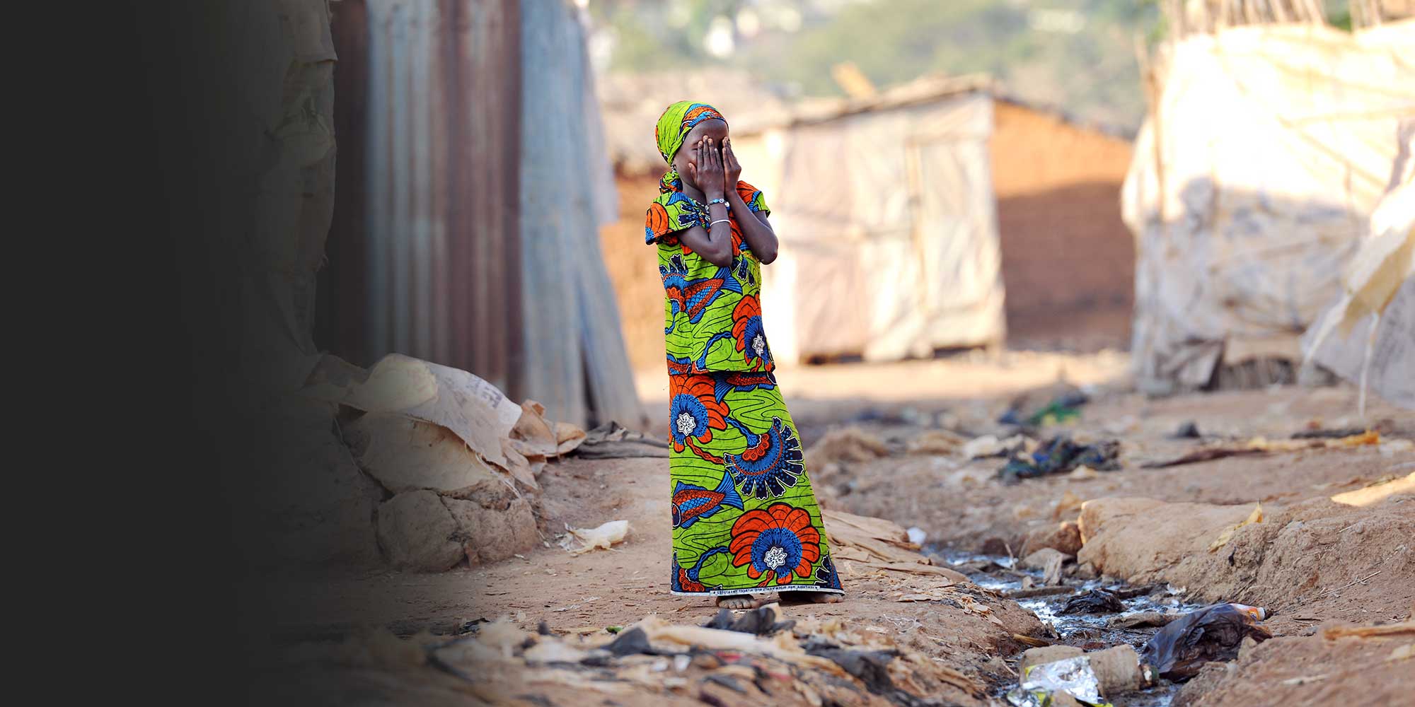 A Nigerian woman in a colourful dress and head wrap stands with her hands over her face. Gavi/Nigeria/Adrian Brooks