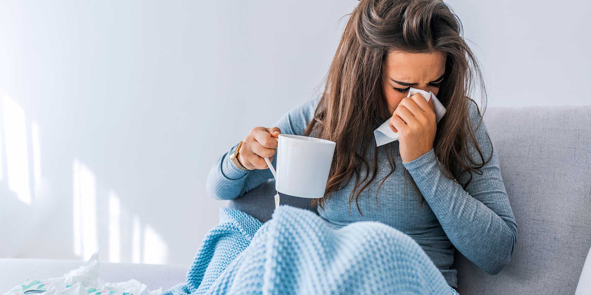 Sick woman with a mug and a tissue over her nose, sitting under a blanket.