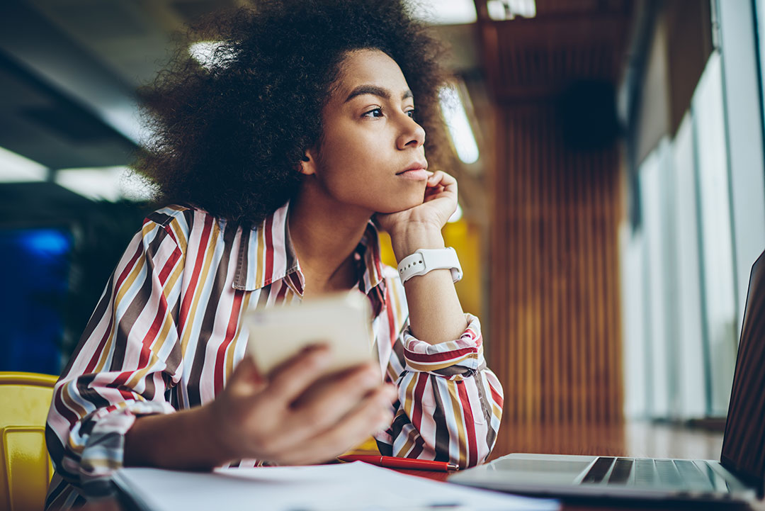 Pondering african american young woman thinking on creative ideas for publication in blog on smartphone device sitting in coffee shop.