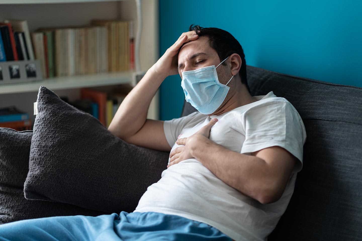 A sick caucasian man wearing face mask feeling sick, headache and cough because of COVID-19 on the sofa in quarantine