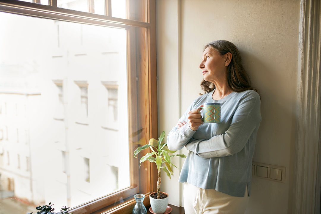 Portrait of thoughtful retired mature woman in casual clothes standing by a window drinking tea.