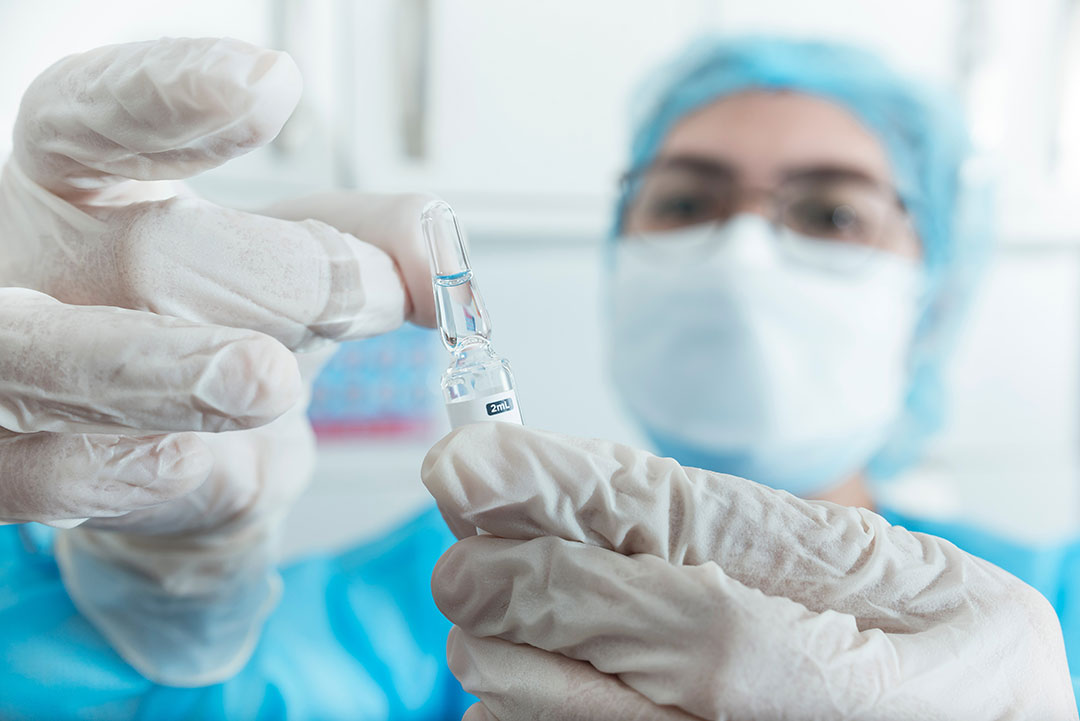 A nurse in protective equipment taps to the head of the ampule to ensure all the liquid drips into the bottom before breaking it.