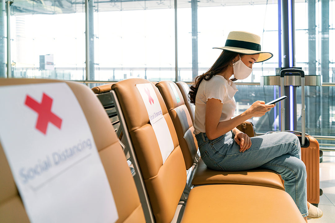 Person wearing mask using mobile phone searching airline flight status and sit social distancing chair in airport