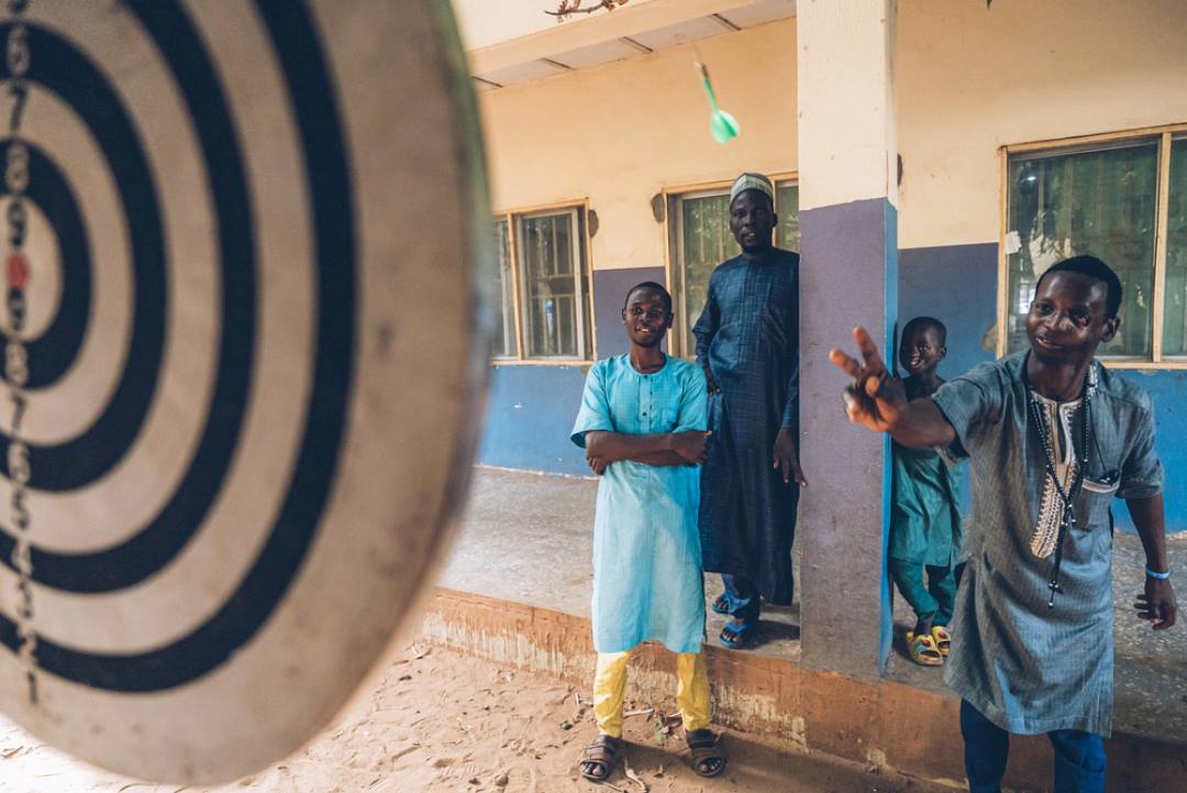 Rabiu, un survivant du noma âgé de 20 ans et originaire de Raba, dans l'État de Sokoto au Nigeria, joue aux fléchettes dans la cour de l'Hôpital Noma de Sokoto. "C'est la première fois que je viens dans cet hôpital. Au début, j'ai refusé, mais ma grande sœur m'a poussé et m'a amené ici", explique-t-il. Il est venu avec son frère Bello, qui se souvient : "Il a commencé à prendre des remèdes à base de plantes à la maison. Son visage avait un furoncle, et un morceau de métal chauffé a été utilisé pour percer le furoncle. La plaie a été retirée et a guéri. Deux autres sont apparus et la même chose a été faite." Nous sommes le 4 mai 2023.