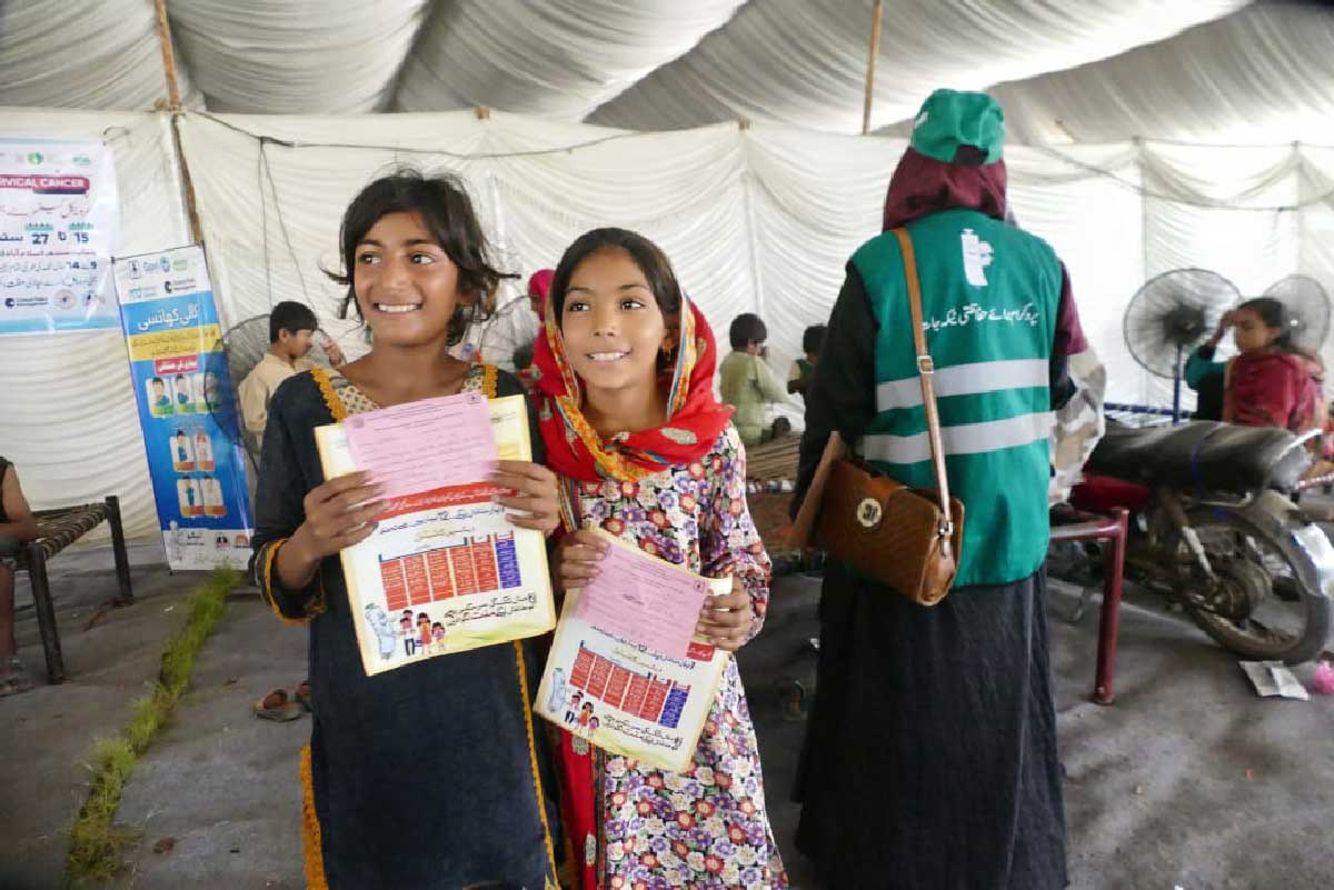 Two smiling girls stand holding their HPV vaccine certificates at a flood relief camp in Punjab district. Credit: Rural Education & Economic Development Society (REEDS)