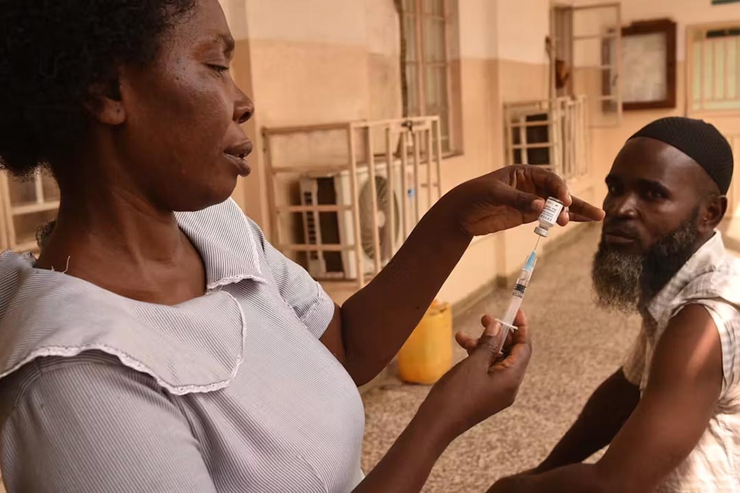 A woman fills a syringe from a small bottle while a man sits with his sleeve rolled up. Credit: Getty Images
