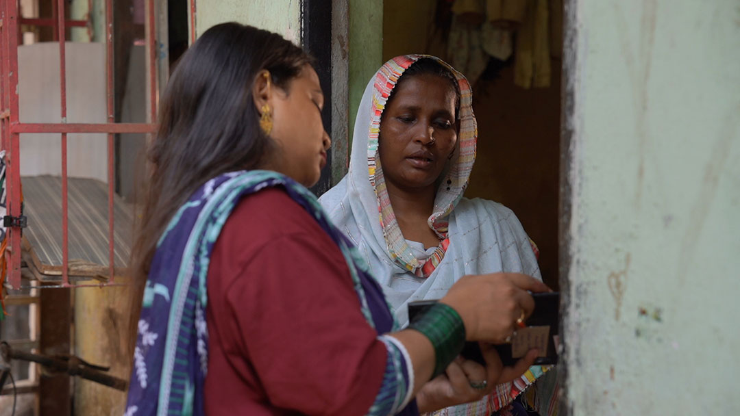 A community outreach worker speaks with a woman at her doorstep, sharing information and recording details. Credit: Amman Shaikh