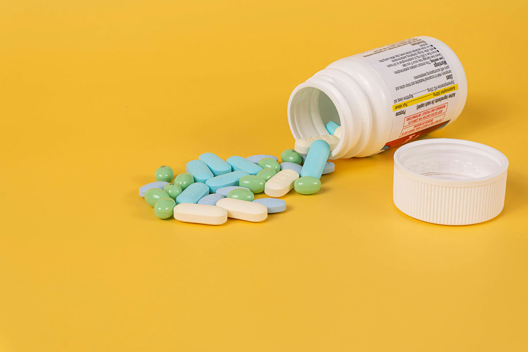 An assortment of pills spilling out of a medication bottle on a pastel orange background. Photo by Etactics Inc on Unsplash