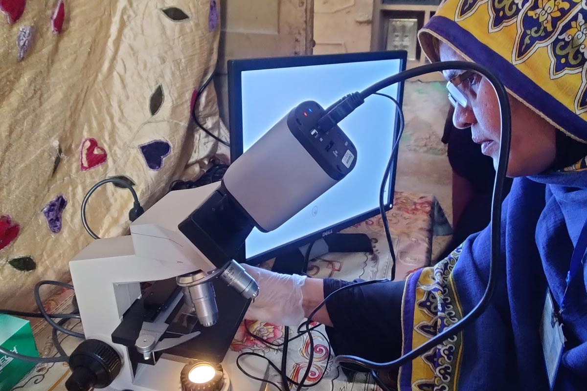 A female health worker looks through a microscope connected to a computer screen, examining a sample during a community health session