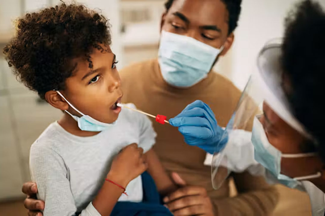 A child receives a swab to test for covid. He is being held by his father, who is wearing a face mask. A female health worker is performing the swab. She is also wearing a face mask. Credit: Drazen Zigic/ Shutterstock